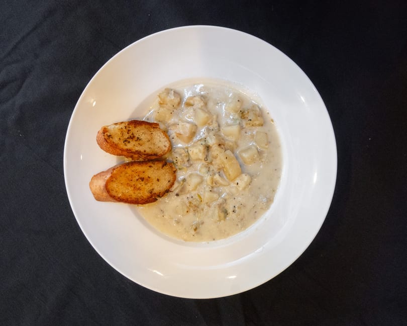 Creamy clam chowder with potatoes and two slices of toasted garlic bread in a white bowl.