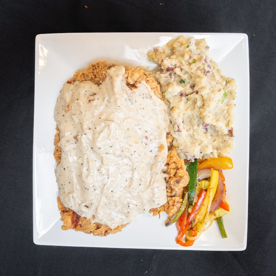 Chicken fried steak with gravy, mashed potatoes, and sautéed vegetables on a white plate.