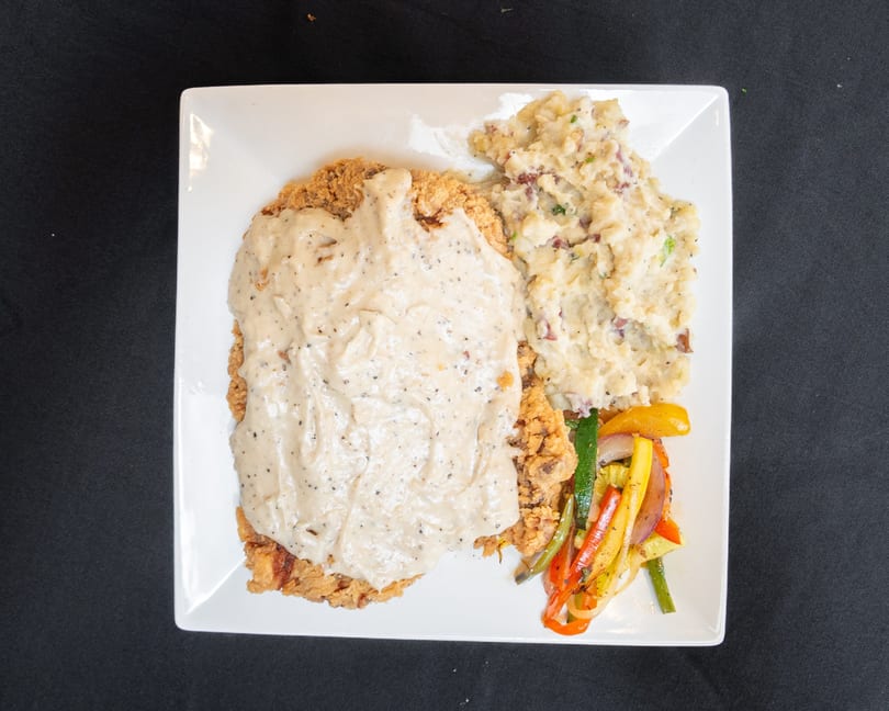Chicken fried steak with gravy, mashed potatoes, and sautéed vegetables on a white plate.