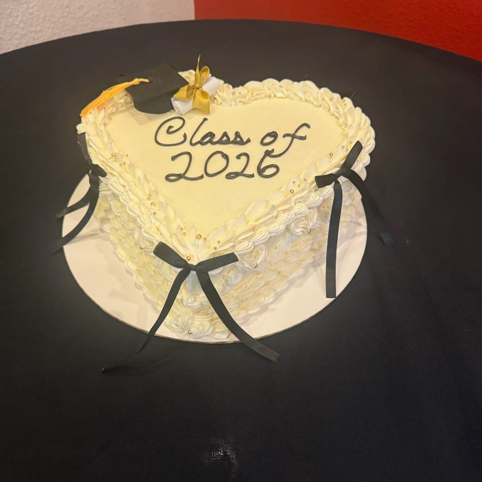 Heart-shaped white cake with "Class of 2026" in black icing, topped with a graduation cap and diploma.