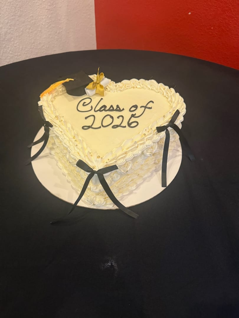 Heart-shaped white cake with "Class of 2026" in black icing, topped with a graduation cap and diploma.