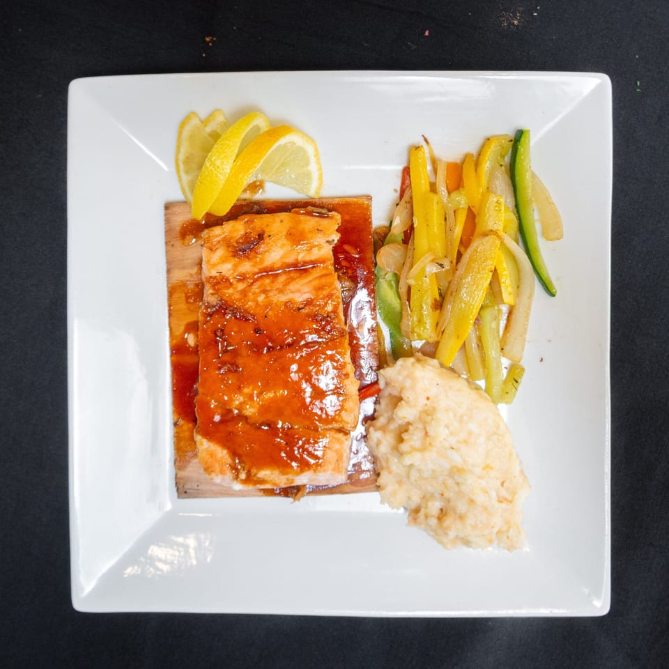 Glazed salmon on a cedar plank with lemon, mashed potatoes, and sautéed vegetables on a white plate.