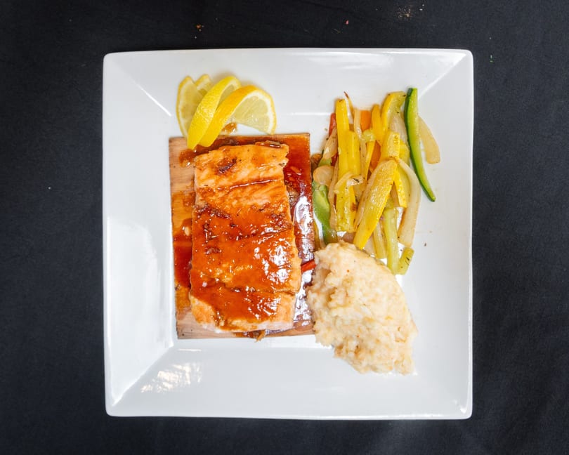 Glazed salmon on a cedar plank with lemon, mashed potatoes, and sautéed vegetables on a white plate.