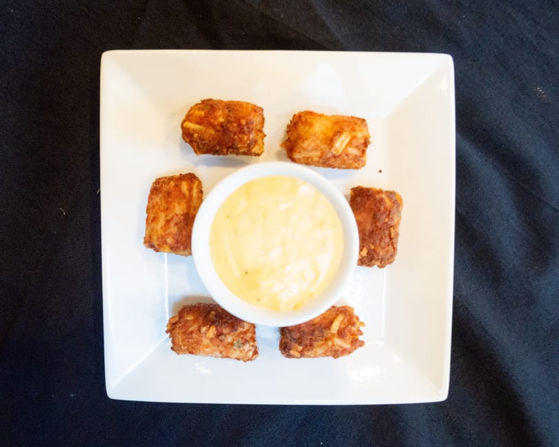 Six golden-brown fried appetizers arranged around a small white bowl of creamy yellow dipping sauce.