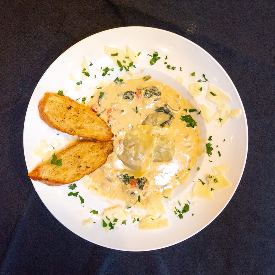 Ravioli in a creamy sauce with spinach and tomatoes, garnished with parsley and parmesan, served with garlic bread.