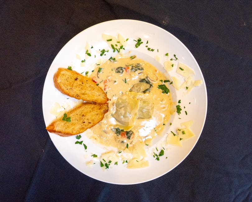 Ravioli in a creamy sauce with spinach and tomatoes, garnished with parsley and parmesan, served with garlic bread.