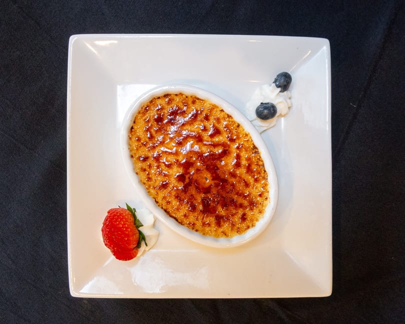 Crème brûlée in an oval dish, garnished with whipped cream, a strawberry, and blueberries.