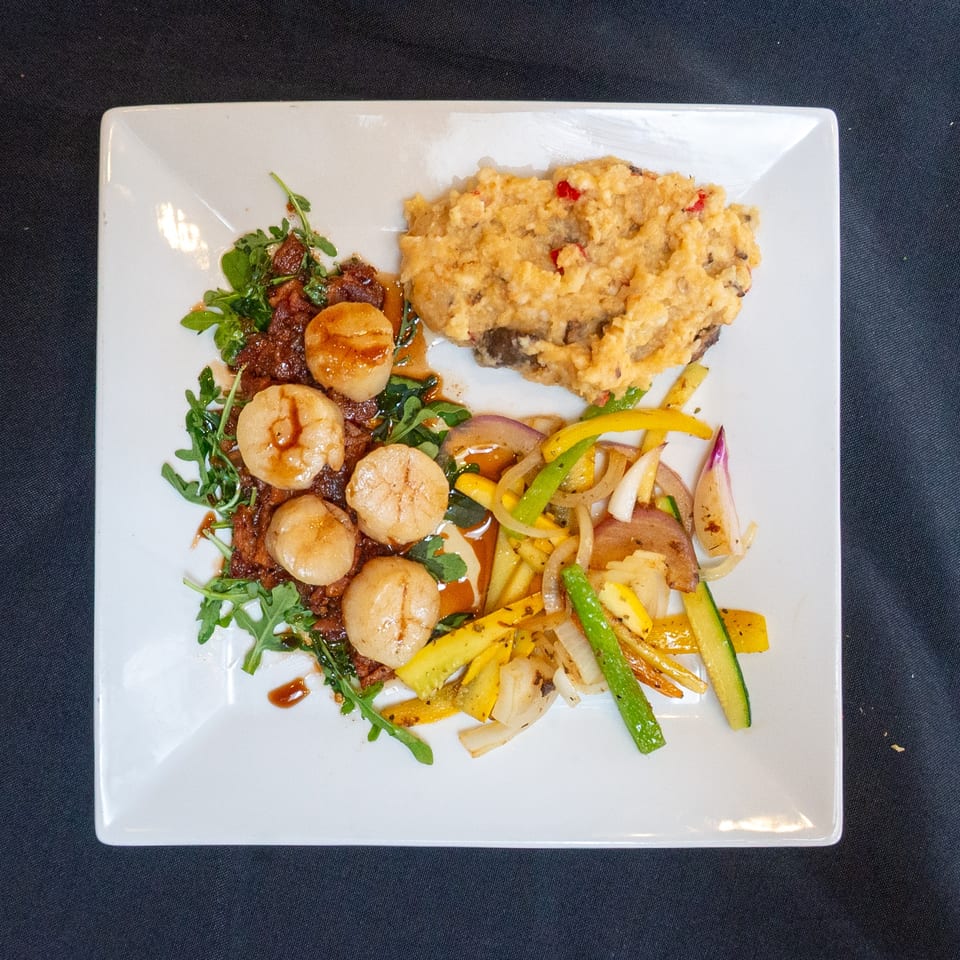 Seared scallops with arugula, mashed potatoes, and sautéed vegetables on a white plate.