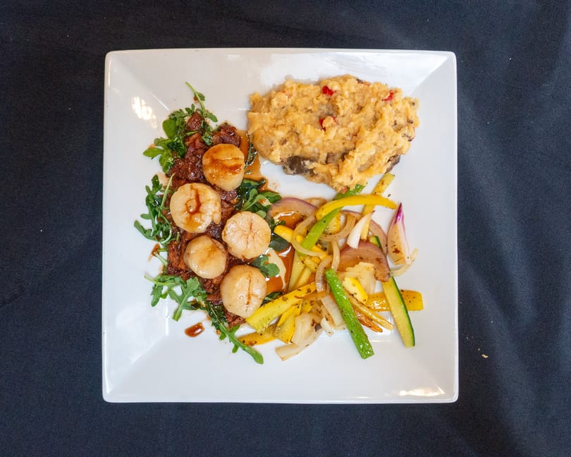 Seared scallops with arugula, mashed potatoes, and sautéed vegetables on a white plate.