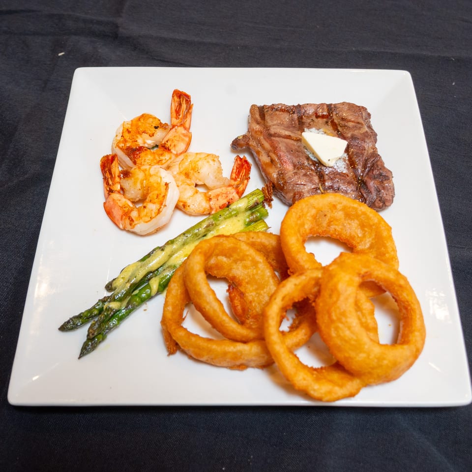 A white plate with steak, shrimp, asparagus, and onion rings on a black tablecloth.