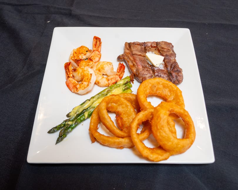 A white plate with steak, shrimp, asparagus, and onion rings on a black tablecloth.