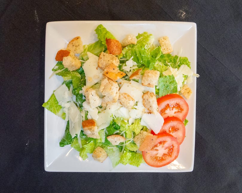 Overhead view of a Caesar salad with romaine, croutons, Parmesan, and tomato slices on a white plate.