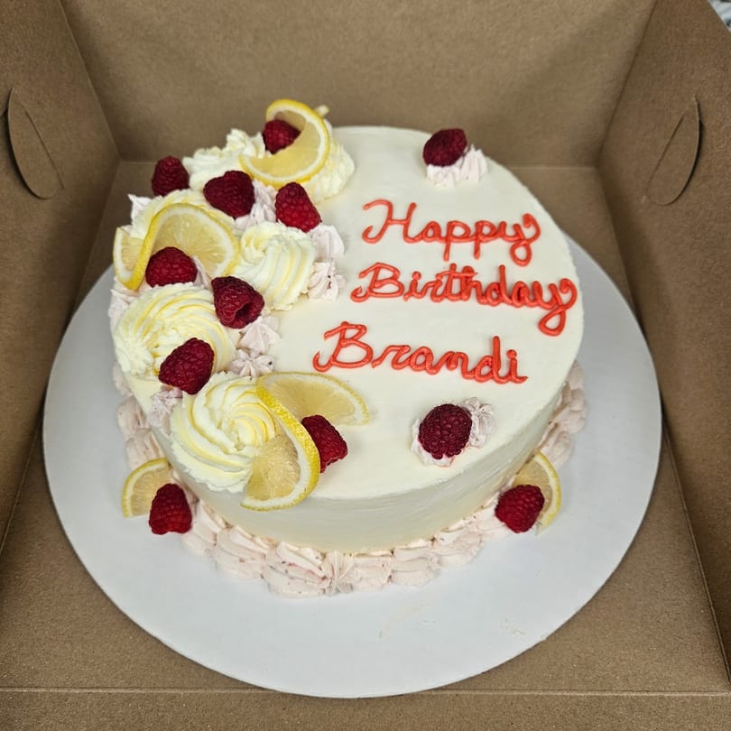 White birthday cake with "Happy Birthday Brandi" in red, topped with raspberries and lemon slices.