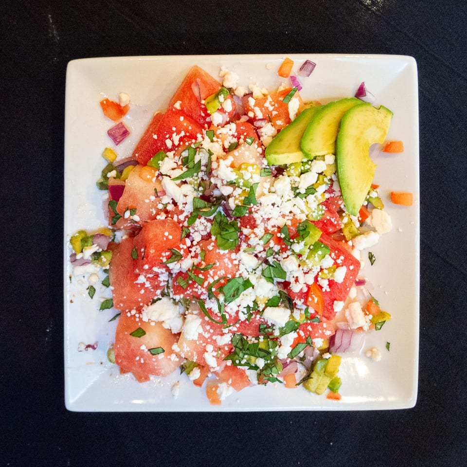 Watermelon salad with feta cheese, avocado slices, and herbs on a white square plate.