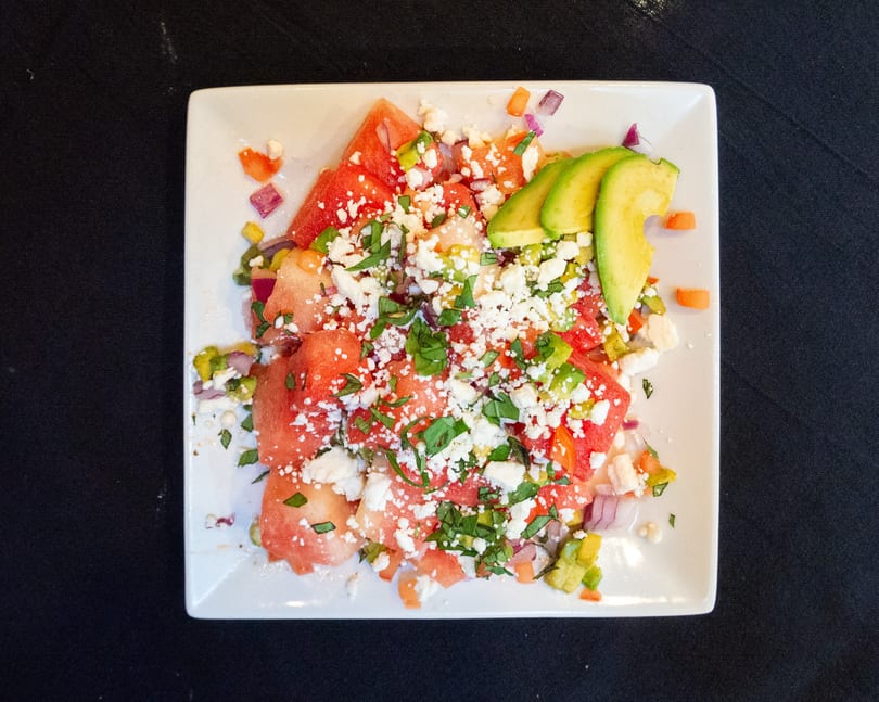 Watermelon salad with feta cheese, avocado slices, and herbs on a white square plate.