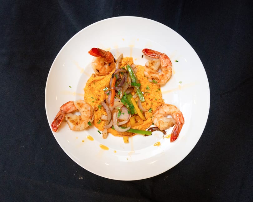 Four grilled shrimp surround a bed of orange puree topped with sautéed onions and peppers.