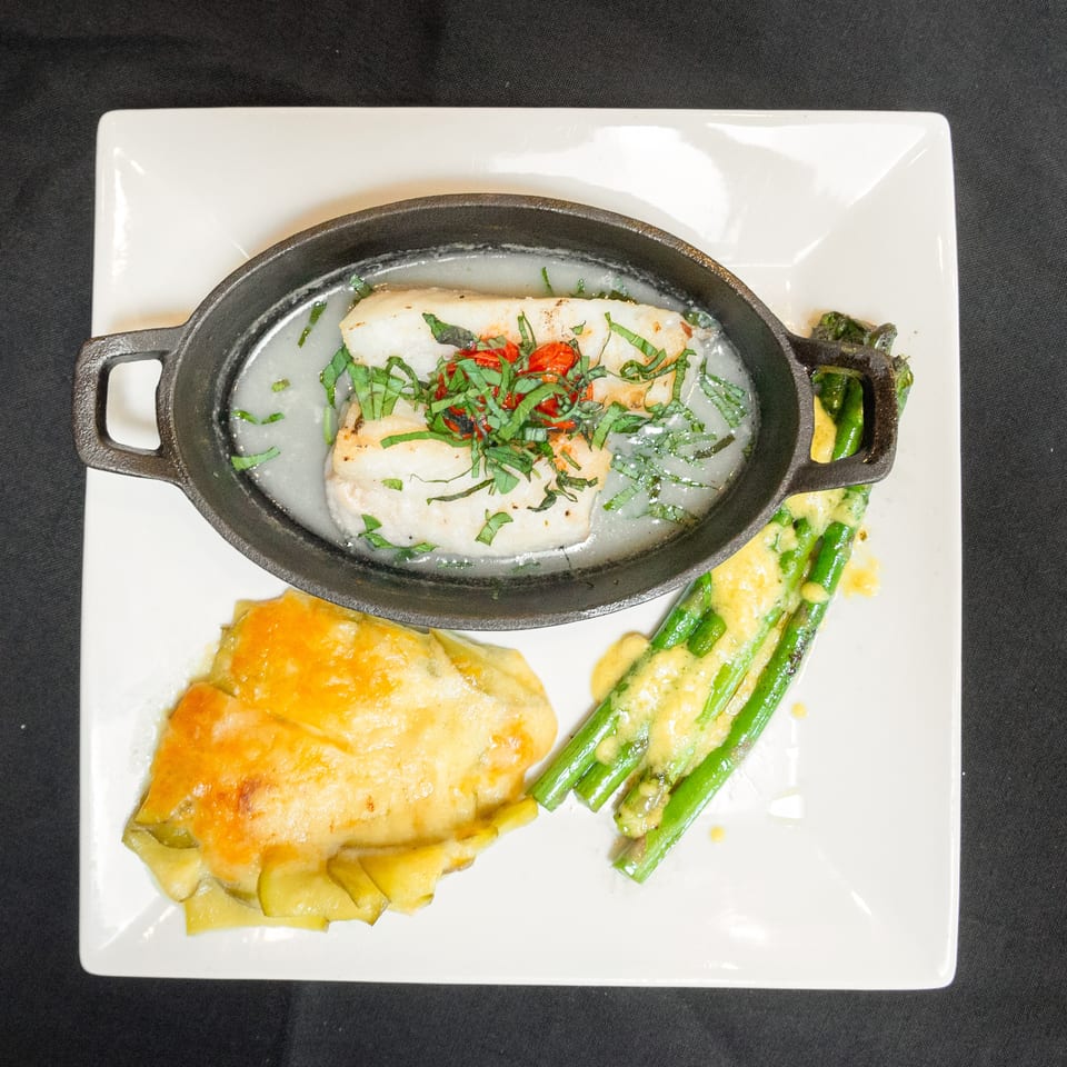 White fish in a cast iron dish with herbs, cheesy potatoes, and asparagus with sauce.
