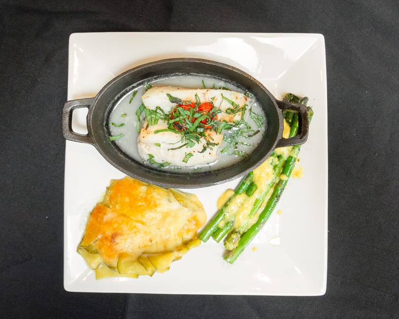 White fish in a cast iron dish with herbs, cheesy potatoes, and asparagus with sauce.