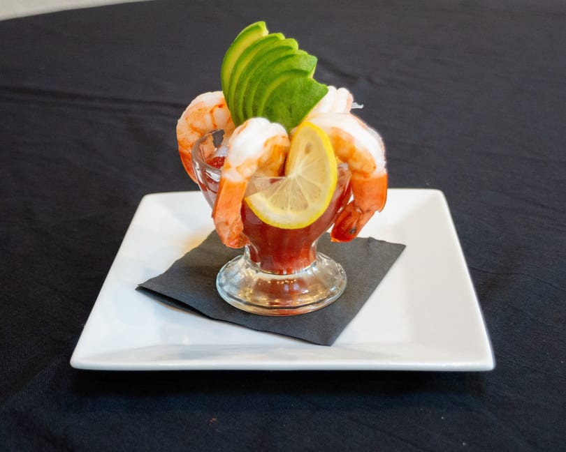 Shrimp cocktail in a glass with avocado and lemon on a white plate with a black napkin.